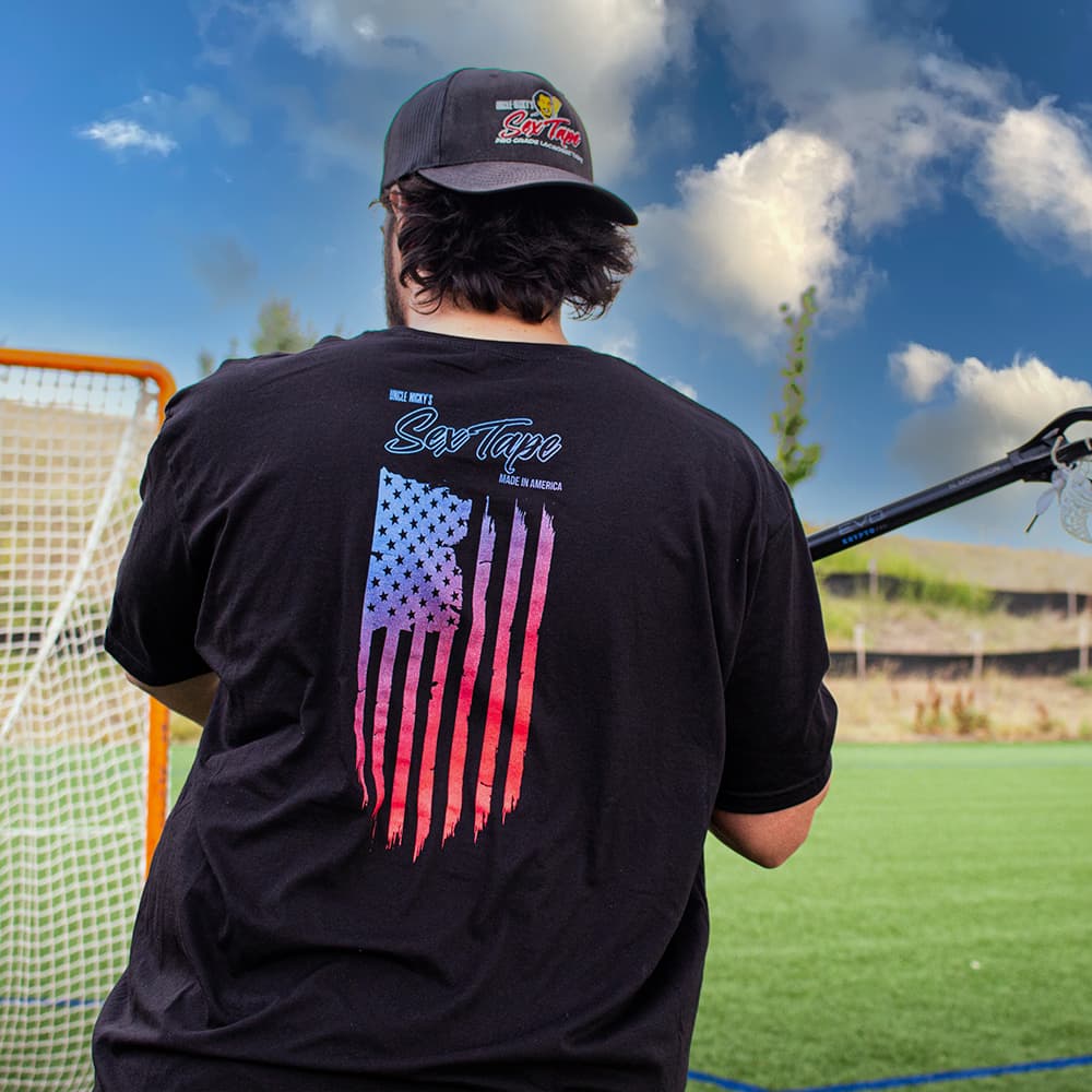 Man holding a lacrosse stick wearing black Uncle Nicky's Sex Tape tee with blue-to-red flag graphic under a blue sky