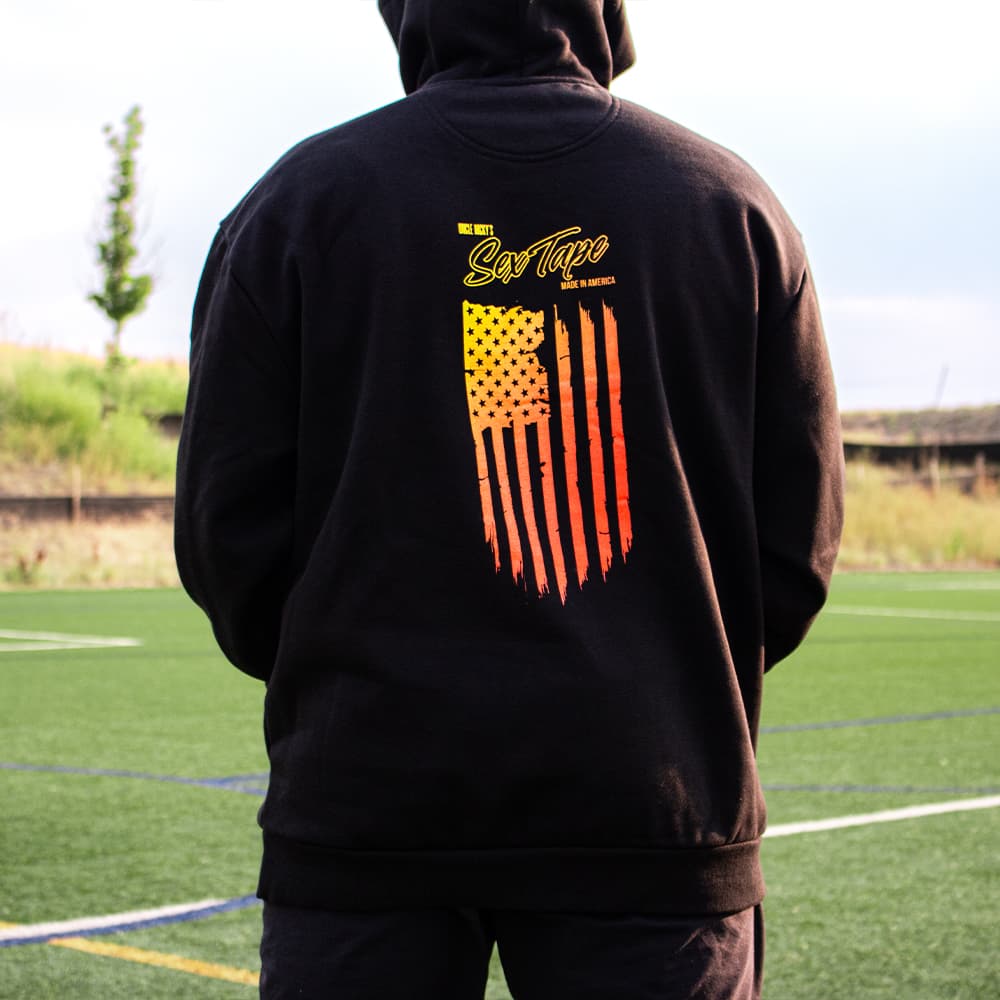 Man wearing black Uncle Nicky's Sex Tape hoodie with yellow-to-red sunset distressed American flag on the back
