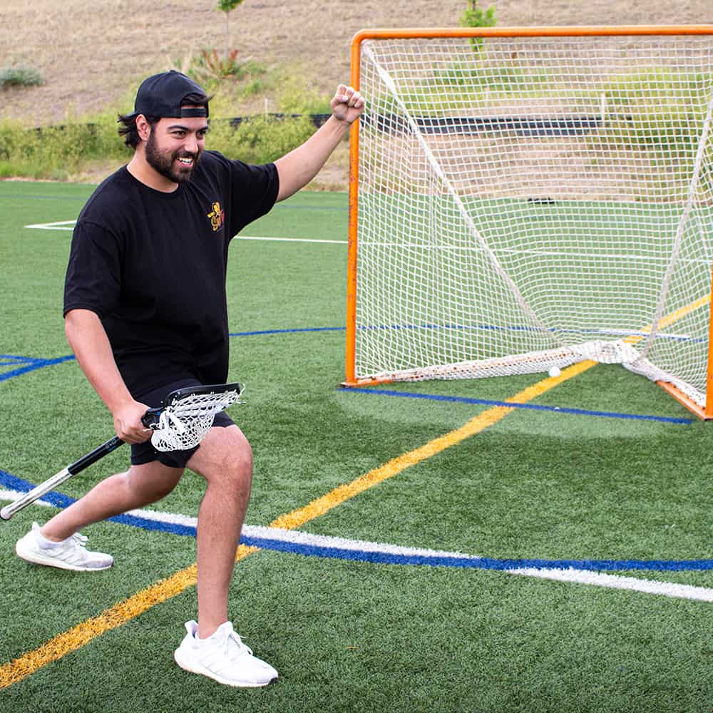 Smiling lacrosse player celebrating with a stick and raised fist, wearing a black Uncle Nicky's Sex Tape tee by an orange goal