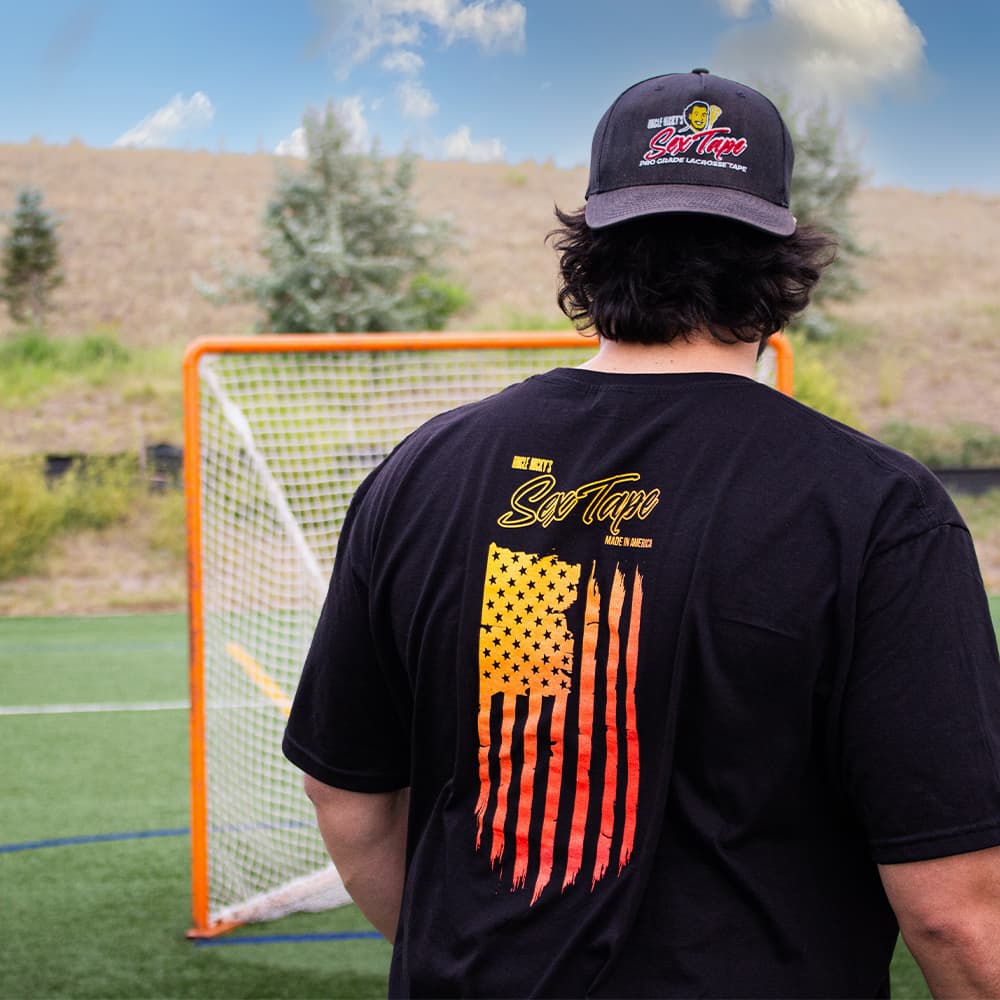 Man wearing black Uncle Nicky's Sex Tape sunset flag t-shirt and logo cap on a lacrosse field by an orange goal